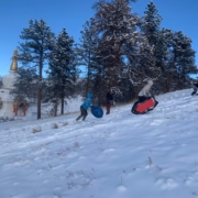 IMG-20250118-WA0004 A photo of three teens and pre-teens running up a snow-covered hillside with sleds. There is a stand of ponderosa pine trees, the Great Stupa of Dharmakaya, and clear blue skies in the background.